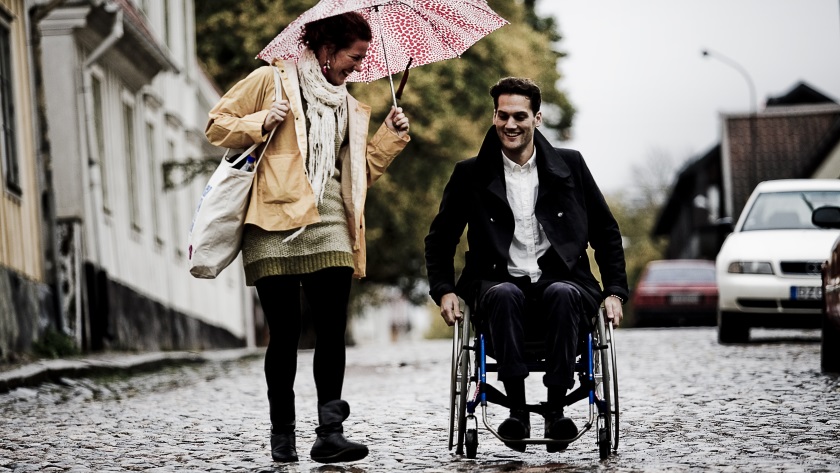 En person sitter i rullstol och kör den framåt längs gatan. En annan person går bredvid pratar, skrattar och håller i ett paraply.