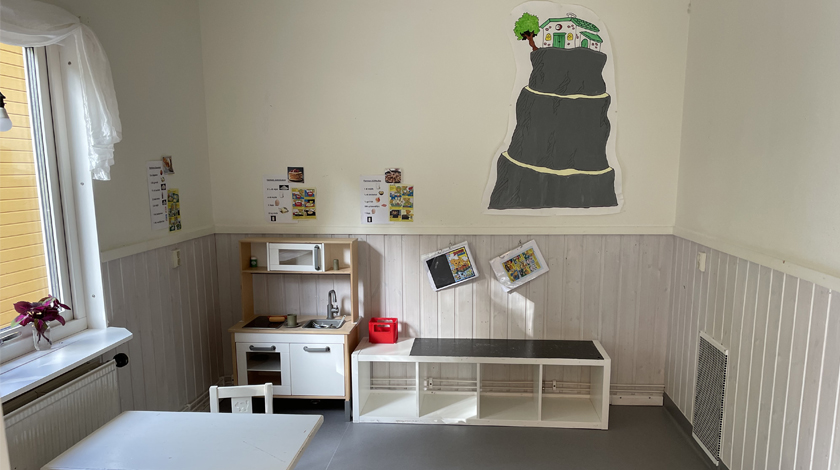 Bild på ett av rummen med stolar och bord, lekkök, hylla och bilder på väggarna